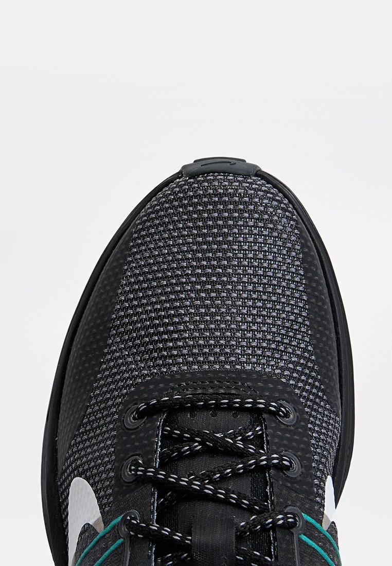 Zapatilla deportiva negra con parte superior de malla texturizada, logo blanco, detalles verdes en los cordones y punta redondeada. Suela de goma para mayor tracción.