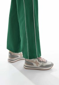 Sneakers in beige and gray with a textured finish, featuring a pink lace and star detail. Paired with green wide-leg pants with white stripes.