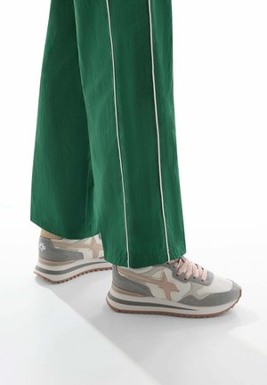 Sneakers in beige and gray with a textured finish, featuring a pink lace and star detail. Paired with green wide-leg pants with white stripes.