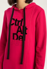 Osoba ubrana w jasnoróżową bluzę z kapturem z czarnymi sznurkami i czarnym wyraźnym napisem "Ctrl Alt Del" z przodu, przyciętą na wysokości szyi i tułowia.