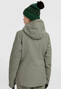 Grijze, gestructureerde skijas met een capuchon, voorzien van een logo op de achterkant en zwarte accenten. Gecombineerd met een groene beanie met een donkere pompon.