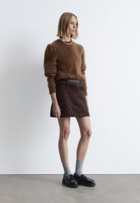 Brauner Mohair-Pullover, tailliert, mit voluminösen Ärmeln. Dunkelbrauter Cordrock, bis zur Mitte des Oberschenkels. Schwarze Slipper und graue Socken runden den Look ab.