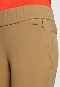 Pantalones deportivos beige con una textura suave, que cuentan con un bolsillo lateral oculto y un detalle sutil de logo cerca de la cintura.