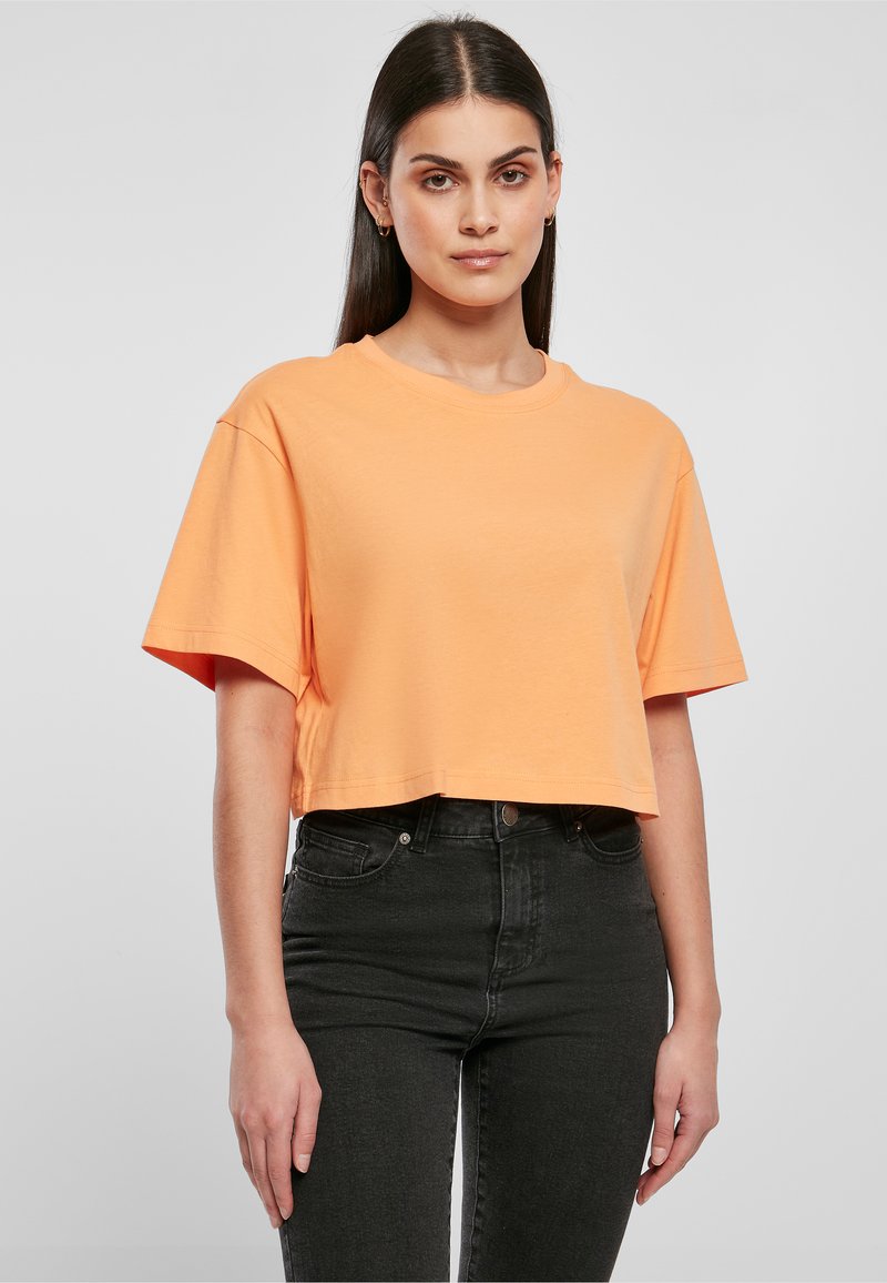 Femme aux cheveux raides foncés portant un t-shirt court orange et un jean noir taille haute sur fond uni.
