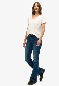 Camiseta de cuello en v de color claro combinada con jeans de campana azul oscuro y zapatos negros. Tejido texturizado, corte casual y cintura con cinturón.