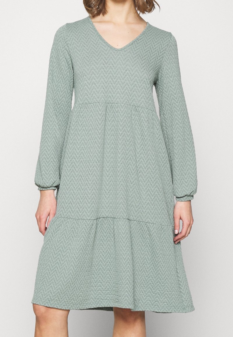 Femme portant une robe texturée vert sauge à manches longues, longueur au genou, avec un décolleté en V et une taille froncée.