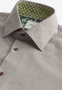 Cămașă de dress texturată, de culoare maronie, cu un guler interior verde cu model. Prezintă nasturi violeți și o etichetă de la "NEXT Tailored."