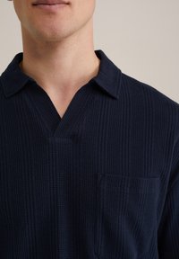 Polo shirt texturat de culoare albastru marin, cu guler, mâneci scurte și un buzunar pe piept cu un design tăiat în diagonală.