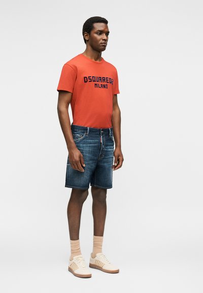 Άνδρας που στέκεται φορώντας πορτοκαλί T-shirt DSQUARED2 Milano, σκούρα τζιν σορτς, μπεζ κάλτσες και λευκά αθλητικά παπούτσια σε απλό φόντο.