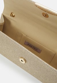 Złota, błyszcząca kopertówka z zapięciem na zatrzask, wyposażona w gładkie wnętrze i wewnętrzną metkę z napisem "ANNA FIELD".