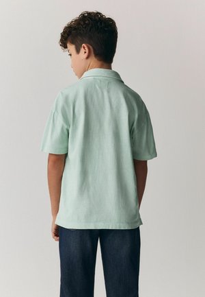 Poika, jolla on kiharat hiukset, pukeutunut vaaleanvihreään lyhythihaiseen paitaan ja tummansinisiin farkkuihin, seisoo kasvojensa suunnassa yksinkertaista vaaleanharmaata seinää vasten.