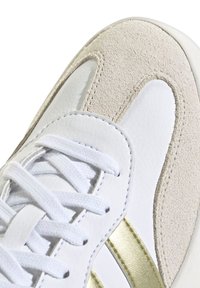Zapatilla blanca de cuero con detalles en ante beige, que cuenta con una franja metálica dorada y cordones blancos. Superficies lisas y texturizadas.