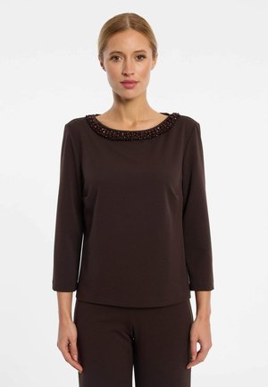 Femme portant un haut à manches longues marron foncé avec un col orné de perles et un pantalon assorti, debout devant un fond blanc uni.