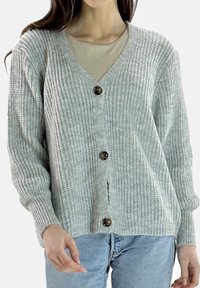 Cardigan grigio chiaro lavorato a maglia con scollo a V, quattro bottoni, trama a coste e maniche lunghe, abbinato a una maglietta beige e jeans blu.