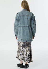 Vrouw met lang blond haar, gekleed in een versleten oversized spijkerjack, een patroon midi-jurk en zwarte chunky sneakers, met haar rug naar de kijker gekeerd.
