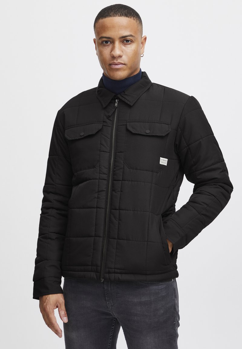 Veste matelassée noire avec zip sur le devant, deux poches poitrine et poches latérales. Dotée d'un col pointu et d'une étiquette en tissu sur le côté gauche.
