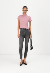 Roze rebrasti kratak top, visoki sivi traperice i crne lakirane cipele na visoku petu. Model nosi malu crnu torbicu. Jednostavna pozadina.