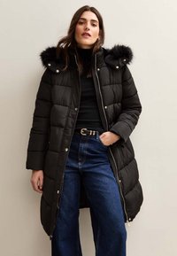 Czarna kurtka puchowa z kapturem z syntetycznym futrem, zapinana na zamek, o pikowanej strukturze. W połączeniu z niebieskimi dżinsami z wysokim stanem.