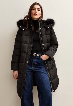 Zwarte pufferjas met een nep-fur capuchon, ritssluiting en gequiltte textuur. Gecombineerd met hoge blauwe jeans.