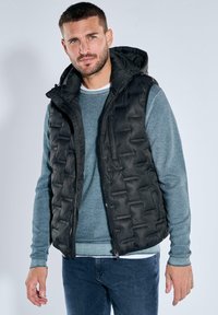 Schwarze Steppweste mit einer strukturierten, gesteppte Optik. Verfügt über eine Kapuze, einen Frontreißverschluss und Seitentaschen, getragen über einem hellblauen Pullover.