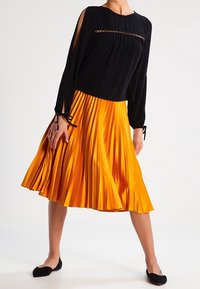 Top nero a maniche lunghe con spalle scoperte e gonna plissettata arancione brillante. La gonna è di lunghezza al ginocchio con una texture lucida; scarpe nere basse.