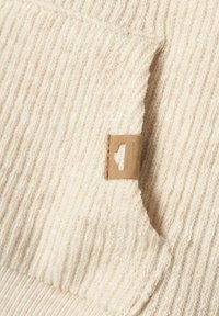 Tessuto a maglia beige con una texture a coste, caratterizzato da un piccolo etichetta rettangolare marrone cucita nella cucitura. Design morbido, leggero e informale.