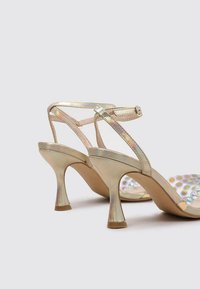 Sandalias de tacón doradas metálicas con un diseño de tiras, que presentan un acabado brillante y un tacón redondeado. Las suelas incluyen una superficie texturizada.