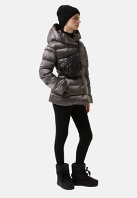 Grå pufferjakke med hætte, sort quiltet taljeveske, sorte leggings og sorte støvler med pelsfor. Glat tekstur og tætsiddende silhuet.