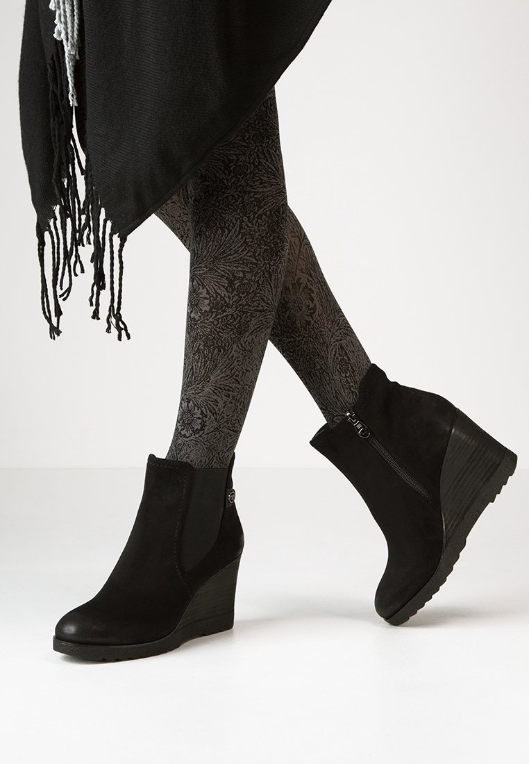 Bottines compensées en daim noir avec une fermeture éclair latérale et un bouton décoratif, associées à des leggings noirs à motifs floraux.