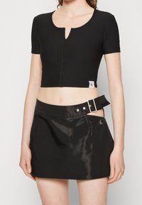 Haut noir court à manches courtes, présentant un décolleté en keyhole et des coutures visibles. Associé à une jupe noire brillante avec un détail de boucles.