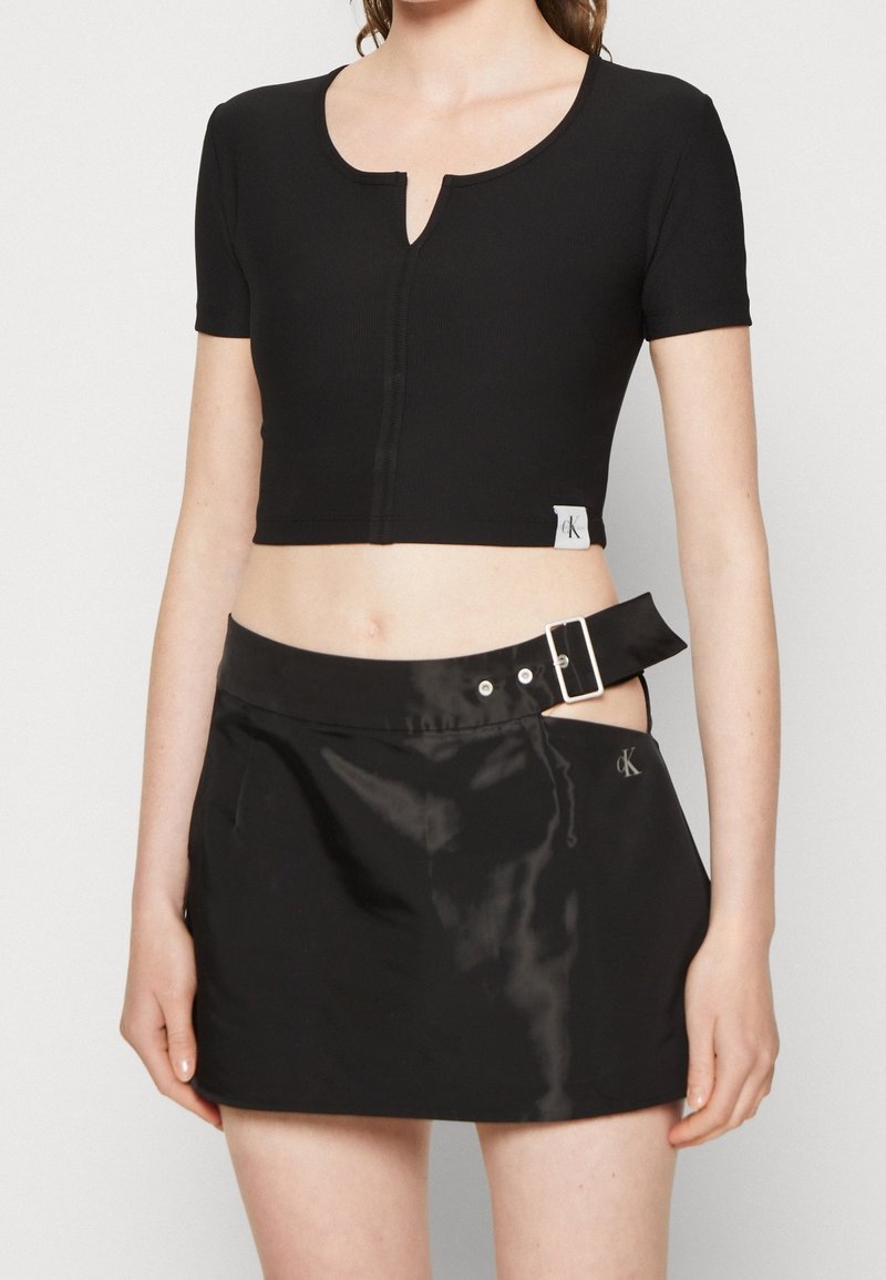 Haut noir court à manches courtes, présentant un décolleté en keyhole et des coutures visibles. Associé à une jupe noire brillante avec un détail de boucles.