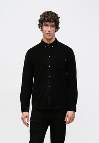 Camicia nera in velluto a coste con colletto alla coreana, caratterizzata da una tasca sul petto e bottoni dorati. Il tessuto presenta un aspetto strutturato e rigato.