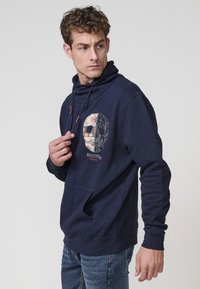 Sudadera azul marino hecha de una mezcla de algodón, con un diseño gráfico de calavera en el pecho, un bolsillo frontal y capucha ajustable con cordón.