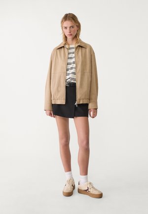 Beżowa zamszowa kurtka z kołnierzem, połączona z paskowanym koszulą i czarną mini spódnicą. Kremowe sneakersy z brązowymi akcentami dopełniają stylizację.