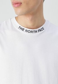 Biała bawełniana koszulka z okrągłym czarnym ściągaczem, na którym widnieje napis "THE NORTH FACE" w pogrubionych wielkich literach. Gładka faktura, prosty design.