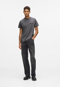 Młody mężczyzna stoi z jedną ręką w kieszeni, ubrany w ciemnoszary T-shirt marki Karl Lagerfeld Jeans, czarne jeansy i czarne buty na białym tle.