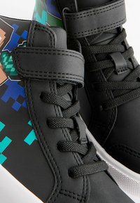 Svarta höga sneakers med en mix av material, snörning, kardborreband och pixelerade grafiker i grönt och blått.