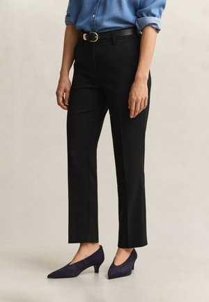 Personne portant un pantalon noir, une chemise bleue à manches longues retroussées, une ceinture noire et des chaussures bleu marine à talons bas sur un fond neutre.
