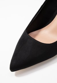 Chaussure à talon haut en daim noir avec un bout pointu, empiècement bas et intérieur beige lisse. Présente un design épuré et minimaliste.