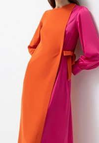 Femme portant une robe bicolore avec un panneau avant orange vif et de longues manches roses, attachée à la taille avec une ceinture orange.