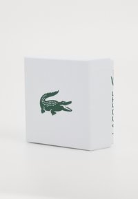 Valge tekstuuriga kast, millel on roheline krokodilli logo ja „LACOSTE” trükituna vertikaalselt roheliselt küljele.