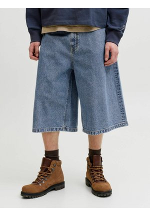 Personne portant un short en denim bleu ample arrivant aux genoux, des chaussures de randonnée marron, des chaussettes foncées et des hauts décontractés superposés.