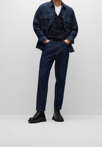 Tmavě modrá džínová bunda s náprsními kapsami, doplněná námořnicky modrou vestou a úzkými tmavými džínami. Oblečení doplňují černé šněrovací boty.