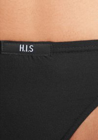 Crne bikini gaće s prošivenim pojasom na kojem se nalazi oznaka "H.I.S." Tekstura je glatka, a tkanina izgleda elastično i tijesno pristaje.