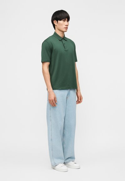Jeune homme debout de profil portant un polo vert foncé, un jean large bleu clair et des baskets blanches sur un fond uni.