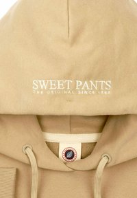 Sweat à capuche beige avec du texte blanc brodé indiquant "SWEET PANTS" et "THE ORIGINAL SINCE 1982." Il dispose de cordons de serrage et de poignets côtelés.