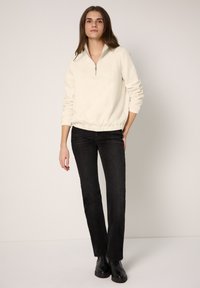 Jasnokremowa bluza rozpinana na zamek z półstojącym kołnierzem, marszczonym dołem i prążkowanymi mankietami, zestawiona z czarnymi spodniami o prostych nogawkach i czarnymi butami.