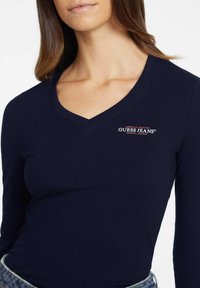 Navyblauw langetermijn T-shirt met V-hals van katoen, voorzien van een klein rood en wit 'Guess Jeans' logo op de linkerborst. Glad textuur.