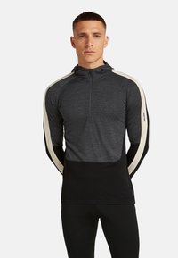 Mand iført en mørkegrå og sort langærmet halv-zip hoodie med cremefarvede striber langs ærmerne, står imod en ensfarvet baggrund.