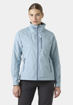 Mujer con chaqueta azul claro con cremallera y cuello alto, con bolsillos con cremallera, combinada con pantalones negros, de pie frente a un fondo liso.